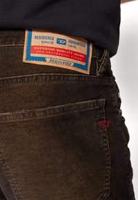 Hnědé kordurové džíny mají na pásku označenou koženou nášivku s modrým a červeným textem, výrazným prošíváním a jemnou texturou.