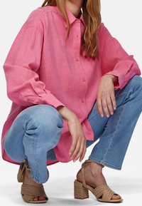 Camicia di lino rosa con bottoni sul davanti e maniche lunghe, abbinata a jeans azzurri e sandali con tacco beige a strappi. Dettagli del tessuto testurizzato visibili.