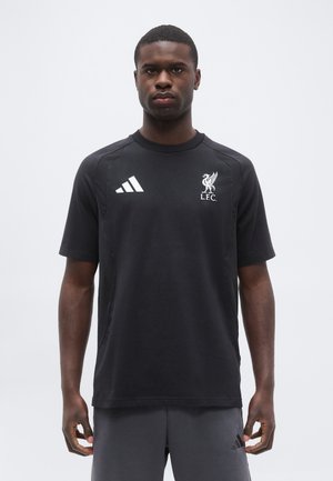 Άνδρας που φοράει μαύρο T-shirt Liverpool FC Adidas και σκούρο σορτς, στέκεται μπροστά σε απλό ανοιχτό φόντο, κοιτώντας μπροστά με ουδέτερη έκφραση.