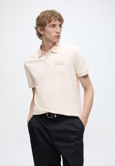 Jeune homme portant un polo beige clair avec le logo "BOSS" et un pantalon noir, debout les mains dans les poches, regardant vers sa gauche.