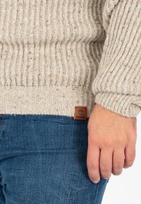 Beżowy sweter prążkowany z teksturowanym wykończeniem i drewnianą naszywką z logo, w połączeniu z niebieskimi dżinsami. Zbliżenie na rękaw i dłoń.