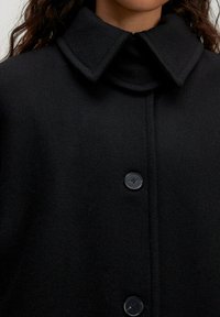 Zwarte wollen blend jas met een hoge kraag, voorzien van een knoopsluiting en een textiele stof. Opvallende details zijn de grote knopen.
