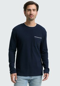 Mann in einem marineblauen Langarmhemd mit Brusttasche und hellblauen Jeans, der vor einem schlichten grauen Hintergrund steht.