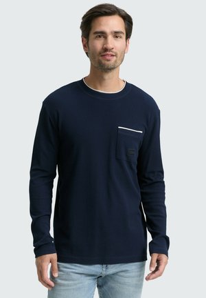 Hombre vistiendo una camisa de manga larga azul marino con un bolsillo en el pecho y pantalones vaqueros de un azul claro, de pie contra un fondo gris liso.