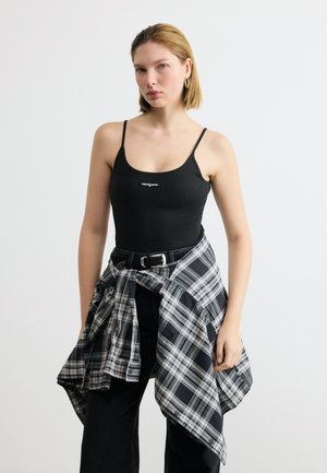 Mlada ženska, oblečena v črno majico brez rokavov z logotipom "tommy jeans", črne hlače in črno-belo karo srajco, zavezano okoli pasu.