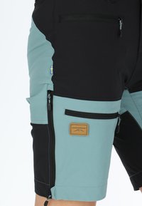 Pantaloni shorts in tessuto nero e teal con multiple tasche con cerniera. Presenta una patch del marchio color sabbia e un piccolo dettaglio bandiera.