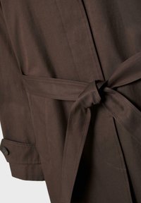 Nahaufnahme des gebundenen Stoffgürtels und des Bündchens mit Knopf eines dunkelbraunen Mantels, die Textur- und Nahtdetails hervorheben.