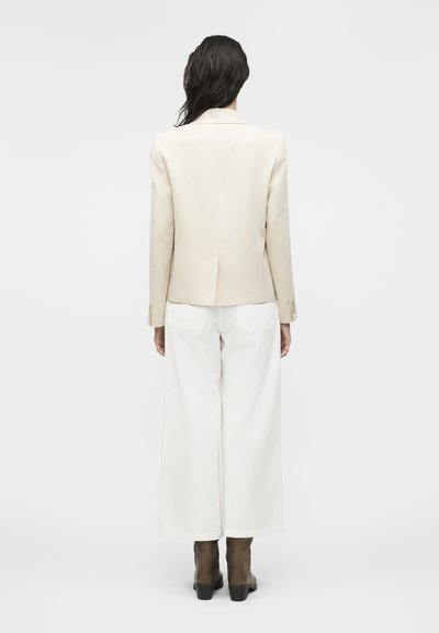 Γυναίκα με βρεγμένα μαλλιά που φοράει μπεζ σακάκι, φαρδιά λευκή παντελόνα και καφέ μποτάκια αστραγάλου, στέκεται με την πλάτη προς το μέρος μας μπροστά σε ένα απλό φόντο.