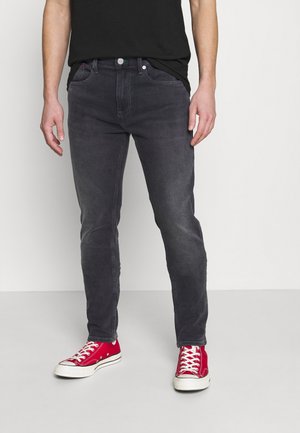 Mand iført mørkegrå slim-fit jeans og knaldrøde høje sneakers, der står mod en ensartet lys baggrund.