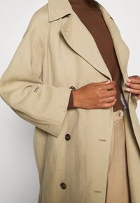 Manteau beige à double boutonnage en tissu texturé, avec de gros boutons, un col cranté et une silhouette structurée. Pull marron en dessous.