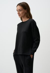 Felpa nera in morbido tessuto, colletto a girocollo, vestibilità rilassata, fessure laterali, indossata con pantaloni neri abbinati, design minimalista, texture liscia.