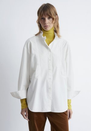 White button-up shirt with a high collar and wide sleeves, layered over a yellow turtleneck. Soft fabric with no visible patterns.
