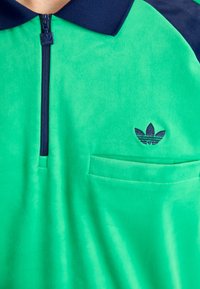 Zelena polo majica od flisa s tamnoplavim ovratnikom, polu-zip zatvaračem sprijeda i malim tamnoplavim Adidas logotipom na prsnom džepu. Mekana tekstura, opušteni kroj.