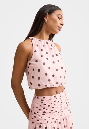 Jeune femme portant un haut rose sans manches et une jupe froncée assortie à pois marron, se tenant devant un fond uni clair.