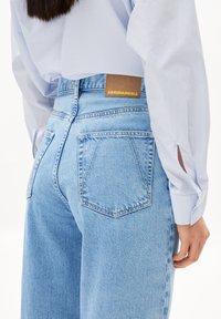 Ljusblå denimjeans med hög midja, som har två bakfickor och en brun läderetikett med "ARMEDANGELS" i guldtext.