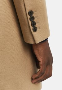 Beige Wollmantelärmel mit vier dunklen Knöpfen, strukturierter Stoff und glatter Oberfläche; teilweise sichtbare Hand, die den Mantel hält.