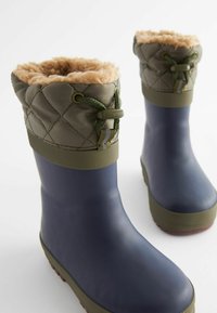 Une paire de bottes en caoutchouc bleu marine avec des poignets en tissu olive vert matelassé et une doublure en polaire beige, conçues pour protéger du froid.