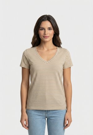 Frau mit langen dunklen Haaren, die ein beiges und weiß gestreiftes T-Shirt mit V-Ausschnitt und kurzen Ärmeln sowie hellblaue Jeans trägt, vor einem schlichten Hintergrund stehend.