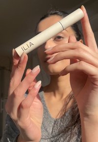 Produkt ILIA w eleganckiej, cylindrycznej tubie w kolorze kremowym, z czarną linią i brandingiem; trzymany w dłoniach z neutralnym lakierem do paznokci.
