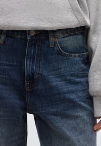 Σκούρο μπλε τζιν με ίσια γραμμή, διακριτικές λεπτομέρειες αποχρωματισμού (whiskering) και πορτοκαλί ραφές. Εφαρμοστή μέση με κλείσιμο με ένα κουμπί και τσέπες.