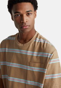 Hnědé tričko s horizontálními pruhy v modré a červené barvě. Má kulatý výstřih a kapsu na hrudi na levé straně. Měkčená tkanina.