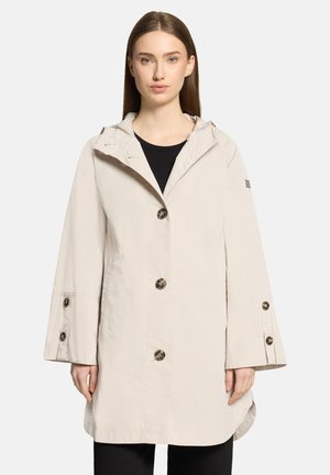 Frau trägt einen beigefarbenen, mit Knöpfen versehenen Kapuzenmantel mit langen Ärmeln über einem schwarzen Oberteil, steht vor einem schlichten hellen Hintergrund.