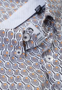 Overhemd met knopen, gemaakt van witte stof met een patroon van tennisrackets en oranje tennisballen. Witte knopen en een contrasterende kraag.