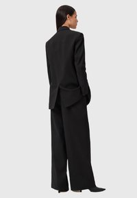 Vrouw in een zwarte oversized blazer en wijde broek, staand in profiel met haar handen in de zakken tegen een effen lichte achtergrond.