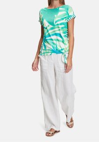 T-shirt with a teal and green tropical leaf print, featuring short sleeves and a knotted detail at the waist, paired with white linen pants.