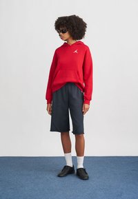 Felpa rossa con cappuccio e tasca a marsupio, abbinata a pantaloni corti blu scuro fino al ginocchio e scarpe da ginnastica nere, con calzini bianchi.