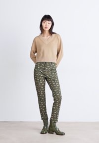 Beige V-halstrui met ribbelzoom, gecombineerd met olijfgroene broek met een hondenprint. Het model draagt groene enkellaarsjes.