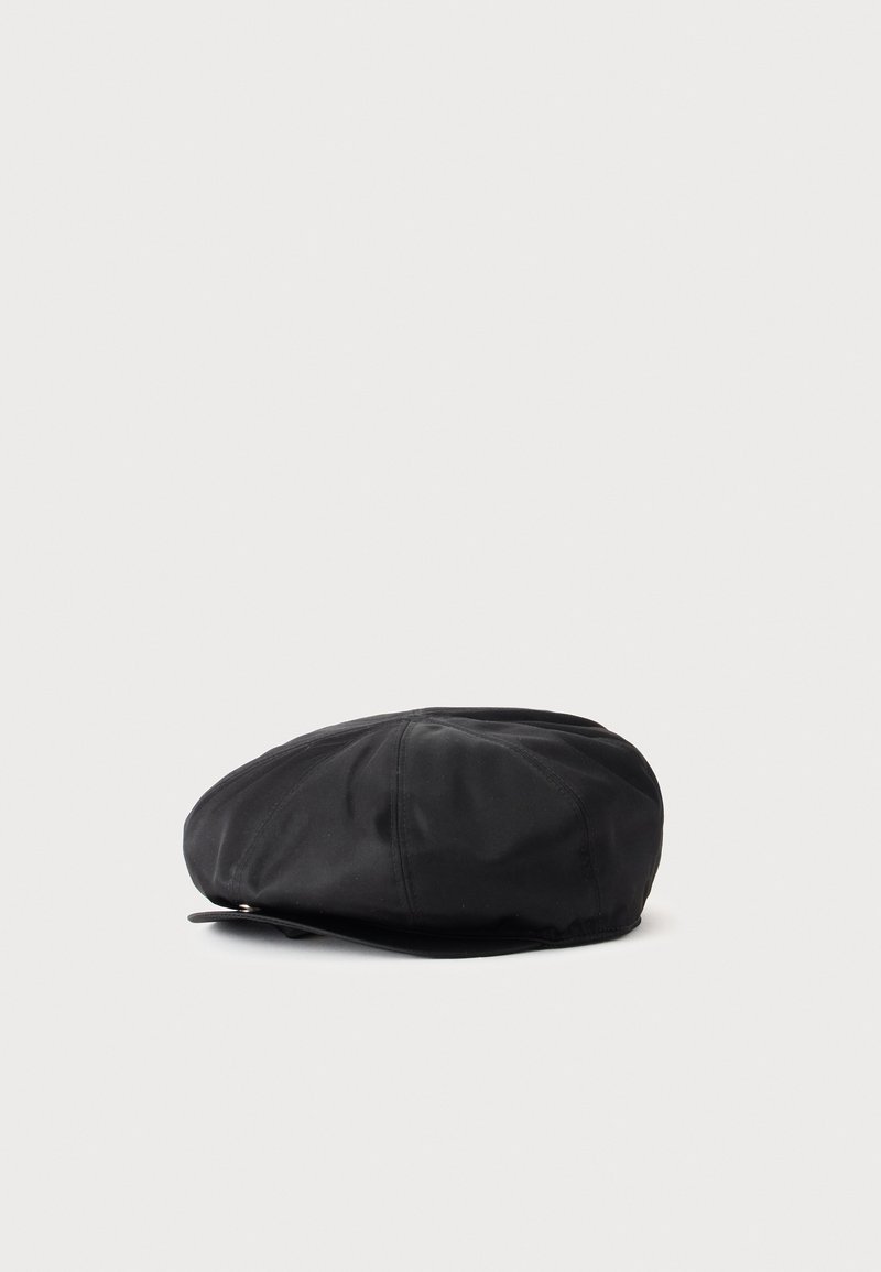Casquette plate noire avec une couronne douce et arrondie et une petite visière, posée à plat sur une surface gris clair.