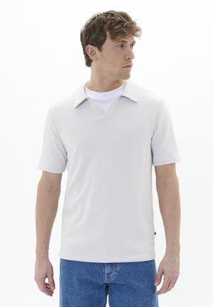 Polo de color gris claro con cuello en V y mangas cortas, hecho de material suave. Lleva puesto una camiseta blanca debajo; combinado con jeans azules.