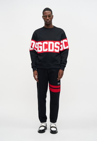 Μαύρη μπλούζα με έντονη λευκή και κόκκινη λογότυπο ταινία στο στήθος, σε συνδυασμό με μαύρες φόρμες που διαθέτουν κόκκινες ρίγες και λεπτομέρεια λογοτύπου.