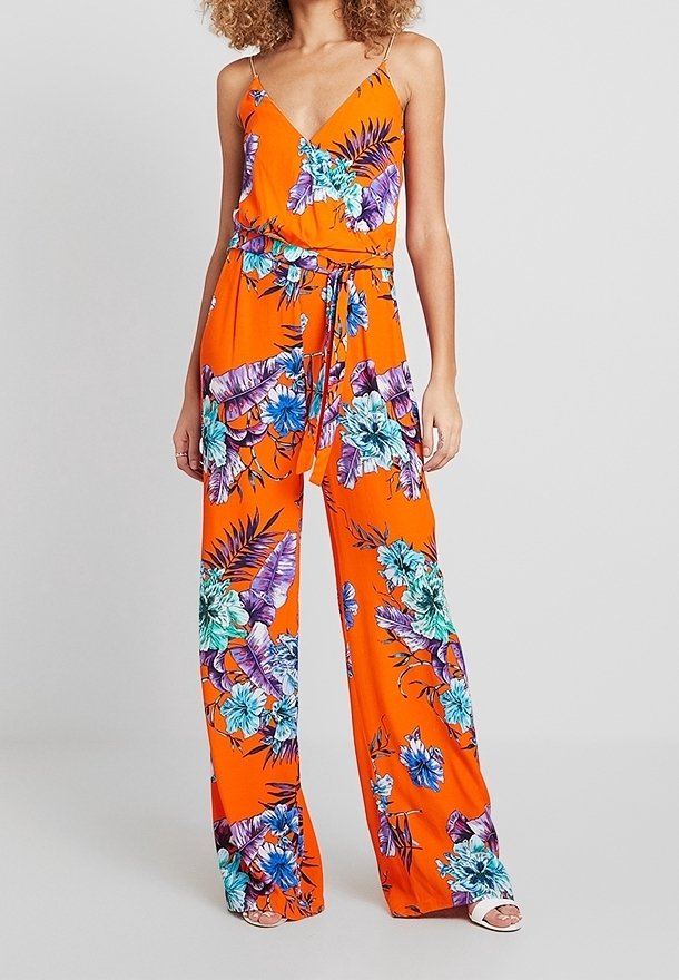 Femme portant une combinaison fleurie sans manches orange avec une taille nouée et un pantalon à jambes larges, debout devant un fond uni.