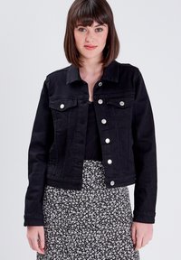 Chaqueta de mezclilla negra con botones en el frente, bolsillos en el pecho y un diseño ajustado. Combinada con una falda de patrón floral en blanco y negro.