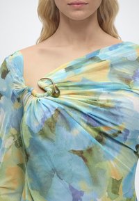 Γυναίκα που φοράει ένα τοπ χωρίς ώμους με παστέλ σχέδιο σε νερομπογιά, με ένα χρυσό δαχτυλίδι και διακοσμητικό πράσινο χάντρα που συγκρ ατεί το ύφασμα κοντά στην κλείδα.