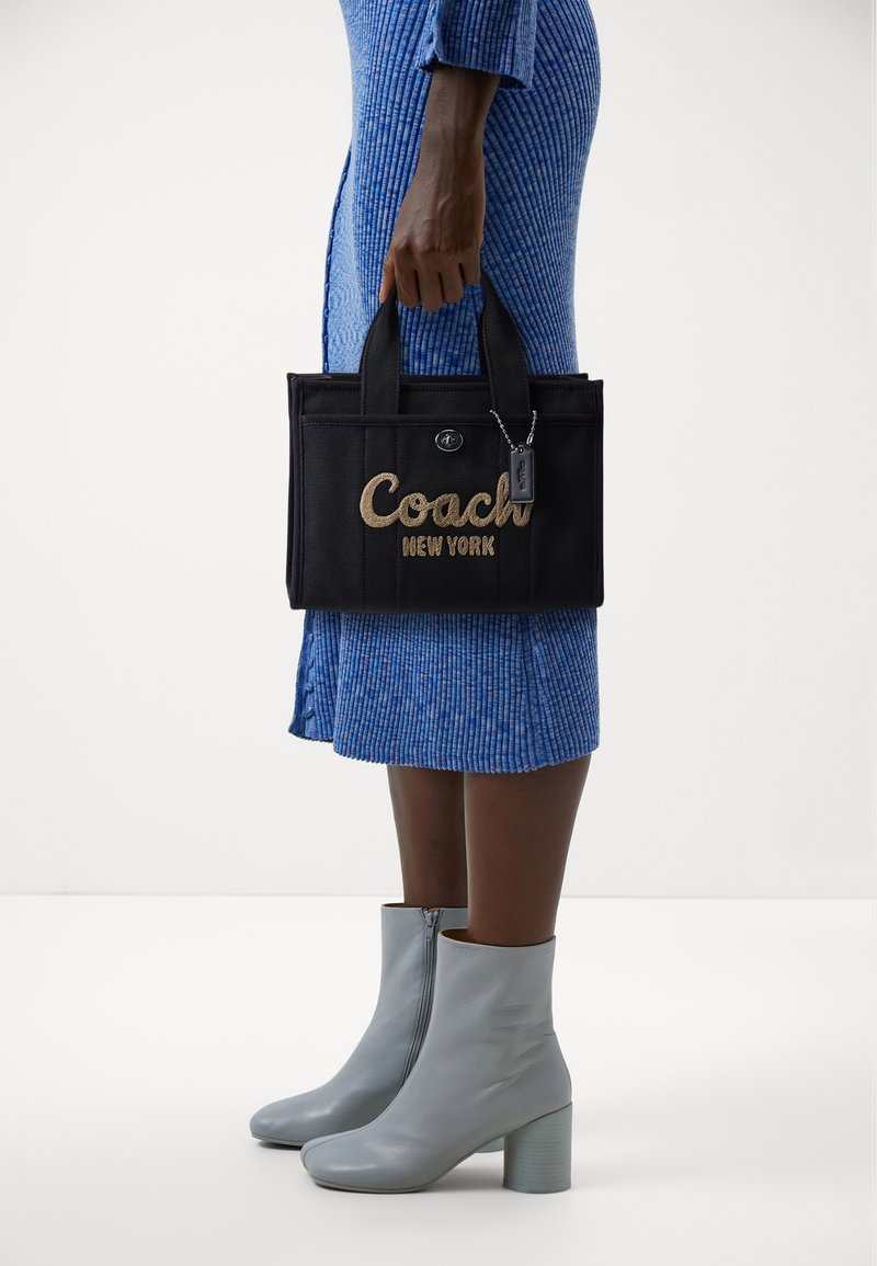 Personne en robe côtelée bleue et bottines gris clair tenant un sac cabas noir Coach New York par les poignées.