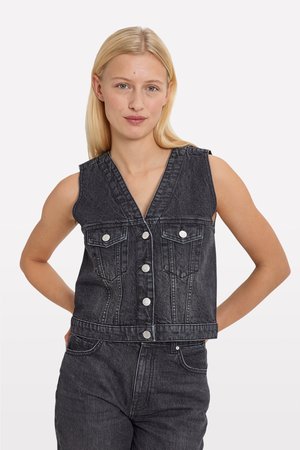 Black denim sleeveless vest with a V-neck, front buttons, two chest pockets, and contrast stitching details. Model stands with hands on hips.