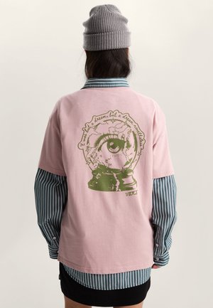 Persona que lleva una camiseta rosa con un gráfico de una bola de nieve que contiene un ojo verde y texto, superpuesta sobre una camisa de manga larga a rayas verdes y un gorro gris.