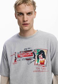 Šedé tričko s krátkym rukávom s farebnou grafikou, ktorá zobrazuje japonské umenie a text, potlačené akcenty a rebrovaný golier.