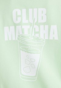 A menta zöld szöveten egy nagy fehér grafika látható, melyen egy jégkockás ital és egy szívószál található, alatta pedig a "CLUB MATCHA" felirat.