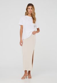 T-shirt bianco in cotone con piccole decorazioni, abbinato a una gonna lunga beige con uno spacco laterale. La modella indossa sandali con tacco beige.