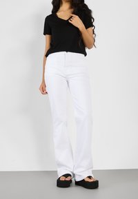 Camiseta negra de manga corta y escote en V combinada con jeans blancos de cintura alta y pantalones acampanados, y sandalias de plataforma negras, sobre un suelo gris.