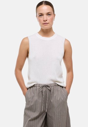 Femme aux cheveux attachés portant un haut en maille blanc sans manches et un pantalon marron rayé taille haute, mains dans les poches.