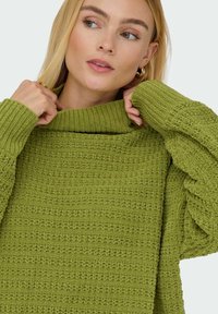 Groene gebreide sweater met een losse col, een textuurpatroon en opgerolde mouwen. De stof lijkt zacht en dik, met opvallende stiksels.