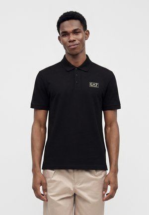 Jeune homme portant un polo noir EA7 Emporio Armani et un pantalon beige, debout devant un fond clair uni.