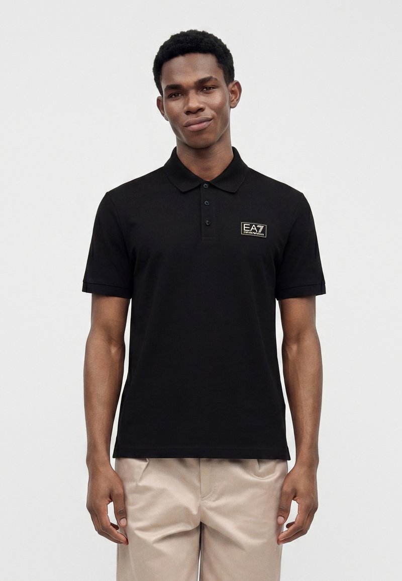 Jeune homme portant un polo noir EA7 Emporio Armani et un pantalon beige, debout devant un fond clair uni.