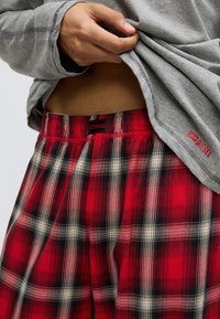 Red and black plaid cotton pajama pants with an elastic waistband and a black label. A gray long-sleeve shirt is partially lifted.