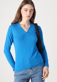 Pulover bleu cu decolteu în V, fabricat dintr-un material moale, având mâneci lungi și un tiv striat. Purtați cu o geantă de umăr neagră texturată.
